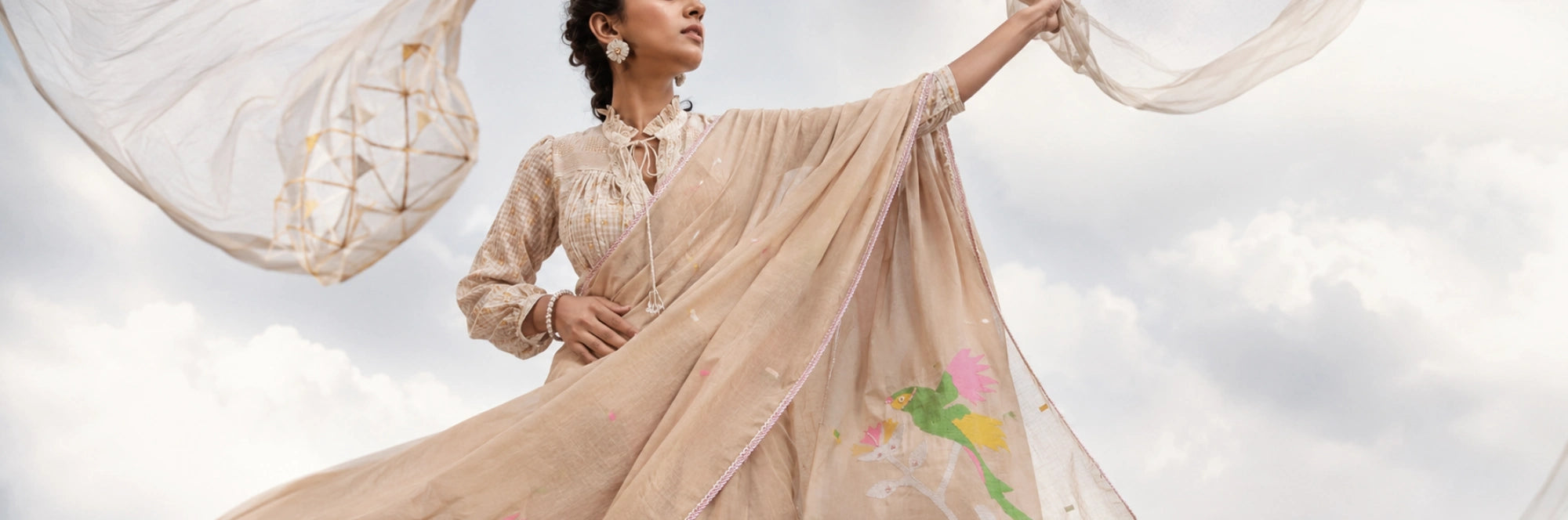 A woman outdoors wearing a flowing beige cotton jamdani  saree  the wind against a cloudy sky